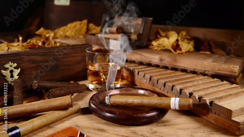 Burning Cigar in an Ashtray with Whisky and Tobacco Leaves on wooden Background
