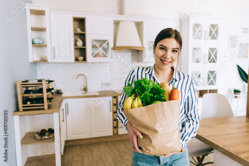 Young smiling caucasian woman hold eco shopping bag with fresh vegetables and baguette in modern kitchen.