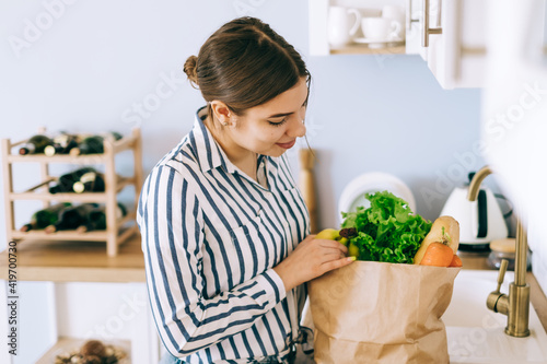 Young caucasian woman taking vegetables from a paper shopping bag in kitchen.