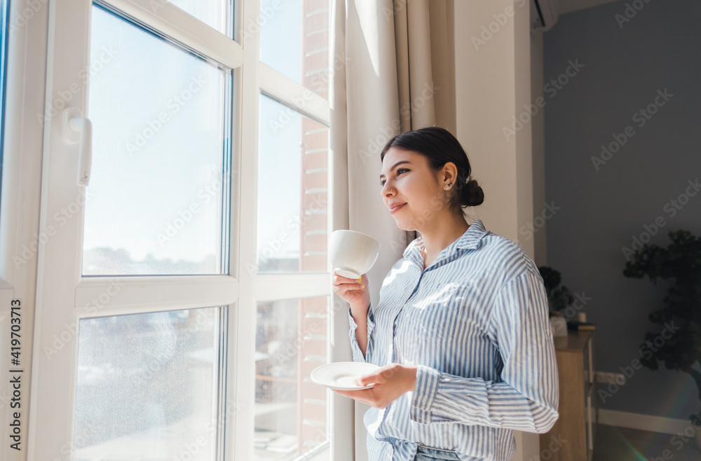 Young smiling caucasian woman standing near the window with a cup of coffee and enjoying the sunny morning. Young smiling caucasian woman standing near the window with a cup of coffee and enjoying the sunny morning.
