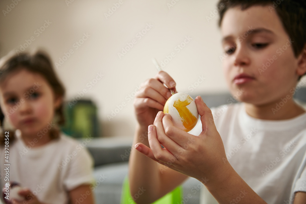 Boy decorating an Easter egg with yellow gouache. Closeup of hands on the background of blurry children Boy decorating an Easter egg with yellow gouache. Closeup of hands on the background of blurry children