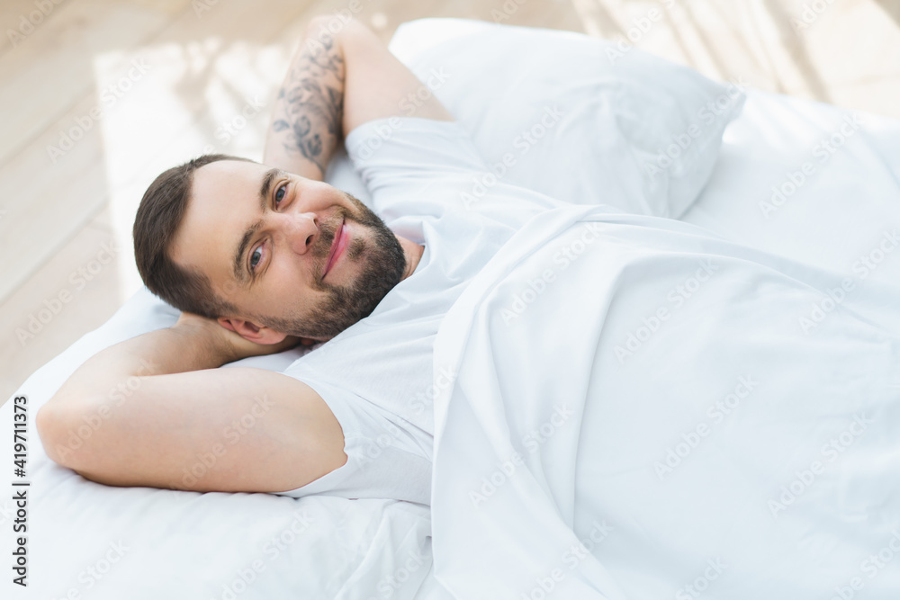 Portrait of handsome young bearded man laying in bed just awake smiling and chilling in the morning enjoying day-off