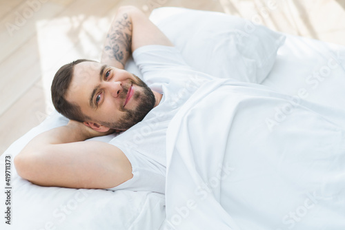 Portrait of handsome young bearded man laying in bed just awake smiling and chilling in the morning enjoying day-off