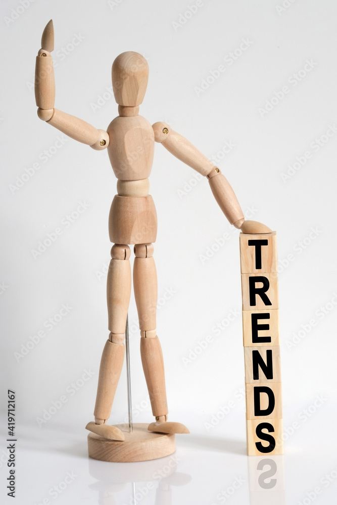 The wooden man raised his hand, and under his left hand he has cubes with the inscription - TRENDS The wooden man raised his hand, and under his left hand he has cubes with the inscription - TRENDS