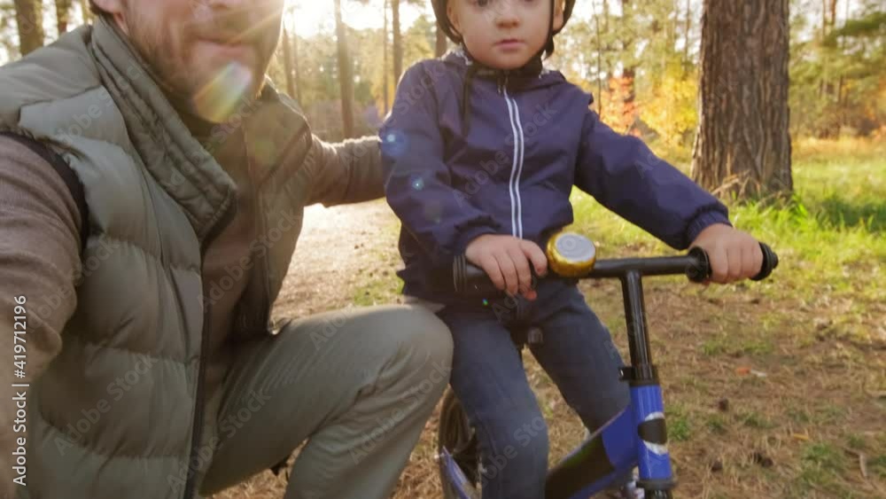 POV shot of father holding camera and filming himself and his cute 5-year-old boy in helmet sitting on balance bicycle in park on sunny summer day POV shot of father holding camera and filming himself and his cute 5-year-old boy in helmet sitting on balance bicycle in park on sunny summer day