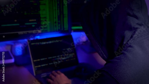 Selective focus on the unidentified man in a hood working as a hacker at the computer in the dark room at night, hacking the system and laundring secret information. photo