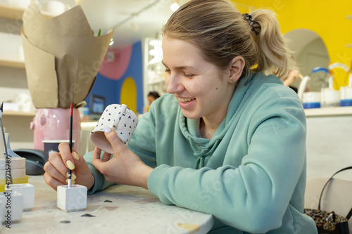 Smiling young woman in casual clothes maks ornament on teecup in a ceramic workshop. Learning new skills, improving skills, hobbies. photo