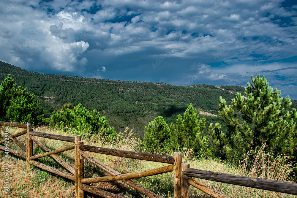 Imagen realizada desde un mirador con vallas de madera y vegetación arbórea en dirección al bosque de montaña y nubes de tormenta