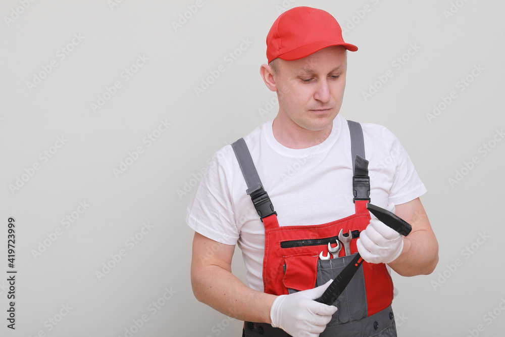 Handsome repairman wearing a red cap and an overall, holding a hammer in his hand, looking seriously, standing on a white background. Maintenance service. building concept. Handsome repairman wearing a red cap and an overall, holding a hammer in his hand, looking seriously, standing on a white background. Maintenance service. building concept.