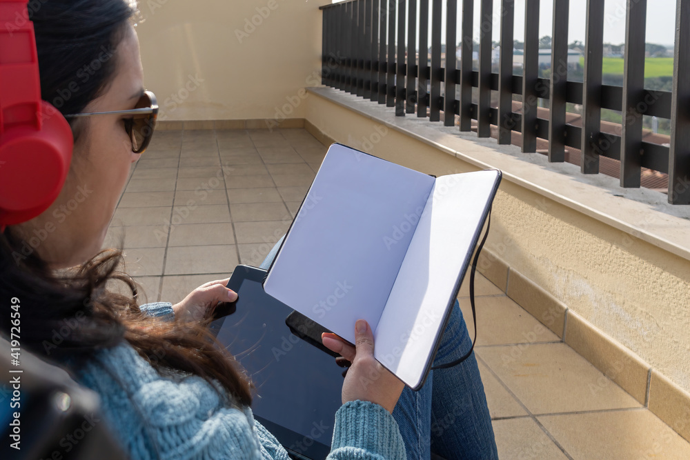 Obraz premium Adult woman sitting in folding chair in her terrace using headphones and looking at empty notebook.