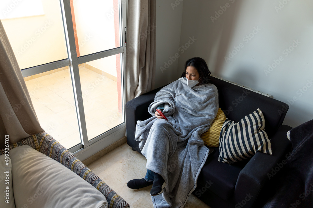 Tired woman using face mask sitting in the couch at home and using mobile phone.