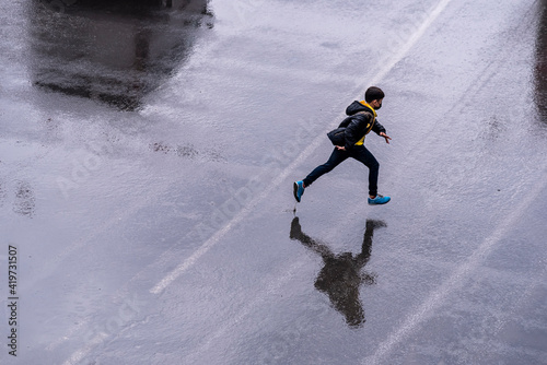 A boy running splashing under rain on street road. top view. Child having fun during rain. Spring outdoor activity for kids. Freedom and happy childhood lifestyle. He wear coat and blue shoes.