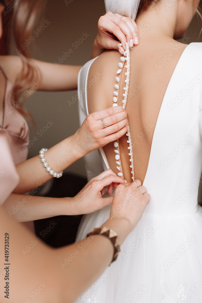 Bridesmaid's hands helping bride fastens with buttons on the back of a beautiful white wedding lace vintage dress close-up, preparation concept in morning for wedding day