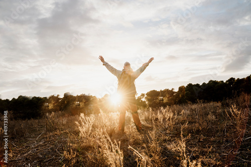 Senior male hiker with arms raised standing in filed during sunset