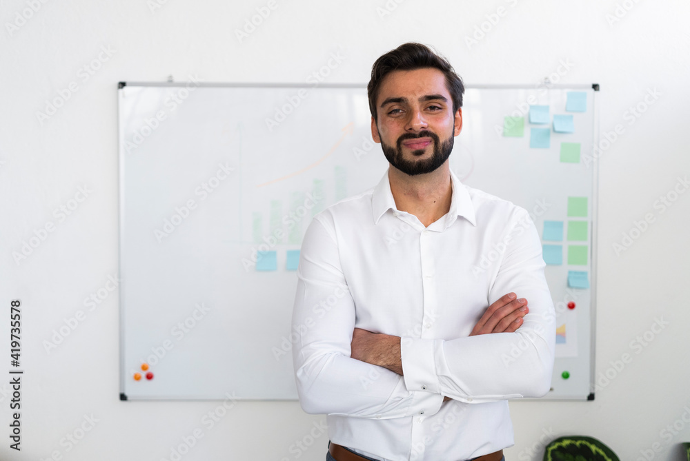 Smiling male entrepreneur with arms crossed standing against whiteboard in office Smiling male entrepreneur with arms crossed standing against whiteboard in office