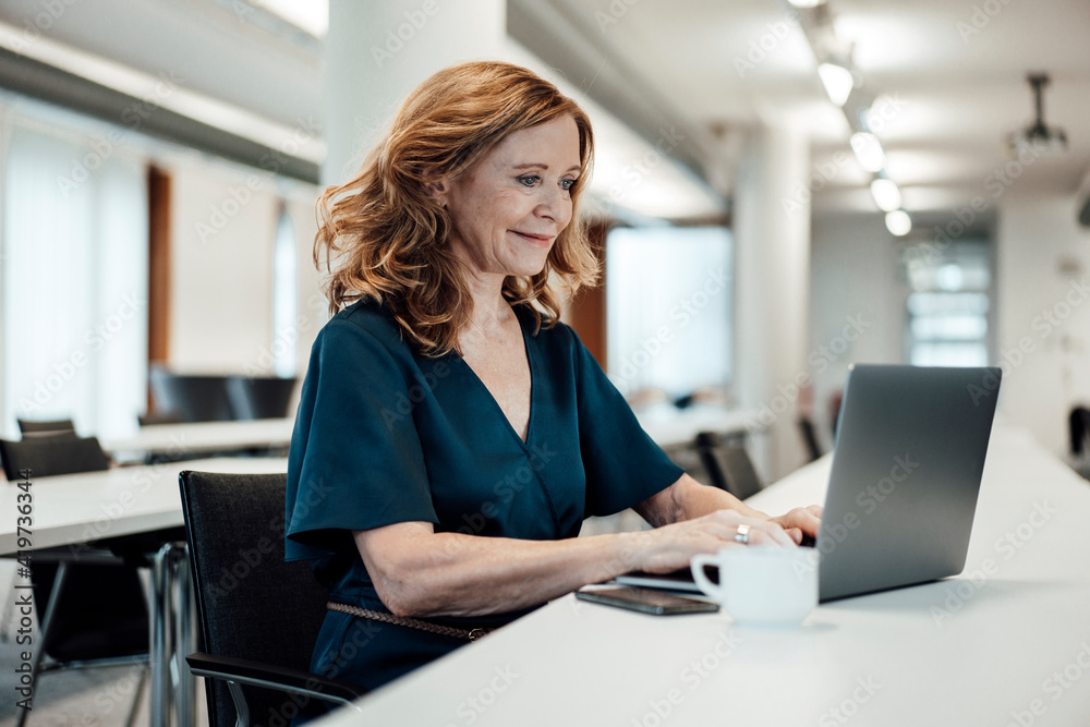 Smiling businesswoman working on laptop at conference table in board room Smiling businesswoman working on laptop at conference table in board room