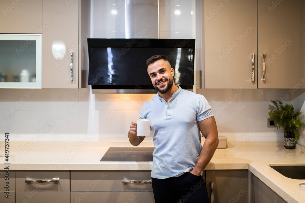 Handsome young man with hands in pockets holding coffee cup while standing in kitchen Handsome young man with hands in pockets holding coffee cup while standing in kitchen