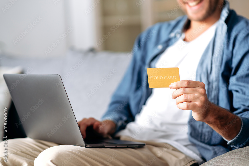 A credit card and laptop in male hands. Joyful caucasian young man in  defocus makes online