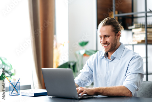 Confident successful guy, freelancer or employee, sits at his work desk at home or in the office, uses a laptop, chatting with colleagues, responding to emails, browsing the Internet