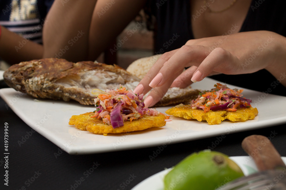 Details of the hands of a woman eating fried fish with patacones, coconut rice and grated carrot salad, cabbage, lettuce. Fried fish is typical in the Caribbean gastronomy of holy week
