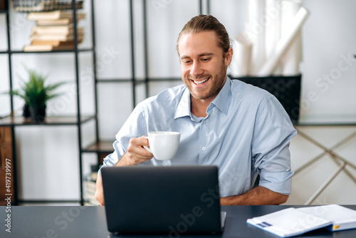 Joyful confident caucasian guy, employee or freelancer, holding a cup of coffee while sitting at his desk at home or office, looking at the camera with a friendly smile © Kateryna