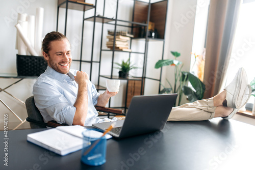 Smiling attractive young man, freelancer or manager, is relaxing at workplace with legs thrown on table. Modern guy uses laptop, browses the Internet, looks for ideas for a project, while sitting at