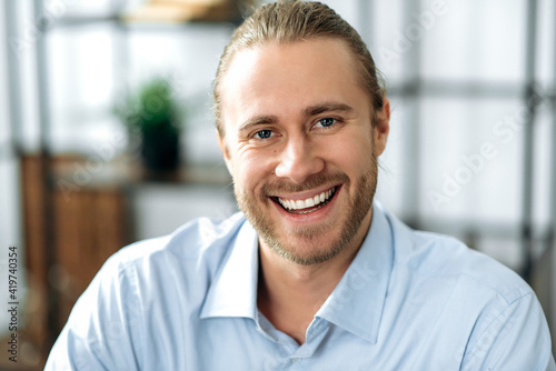 Close up portrait of charismatic handsome confident caucasian guy, wearing formal shirt, sitting at home or office looking at camera with friendly smile