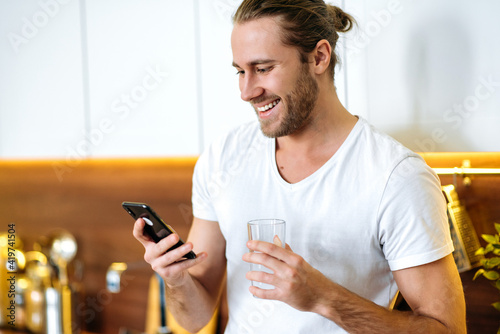 Happy satisfied guy in a white t-shirt, stands at home in the kitchen, holds a glass of clean water, a smartphone in the other hand, chatting online, browsing Internet, answering messages,drinks water