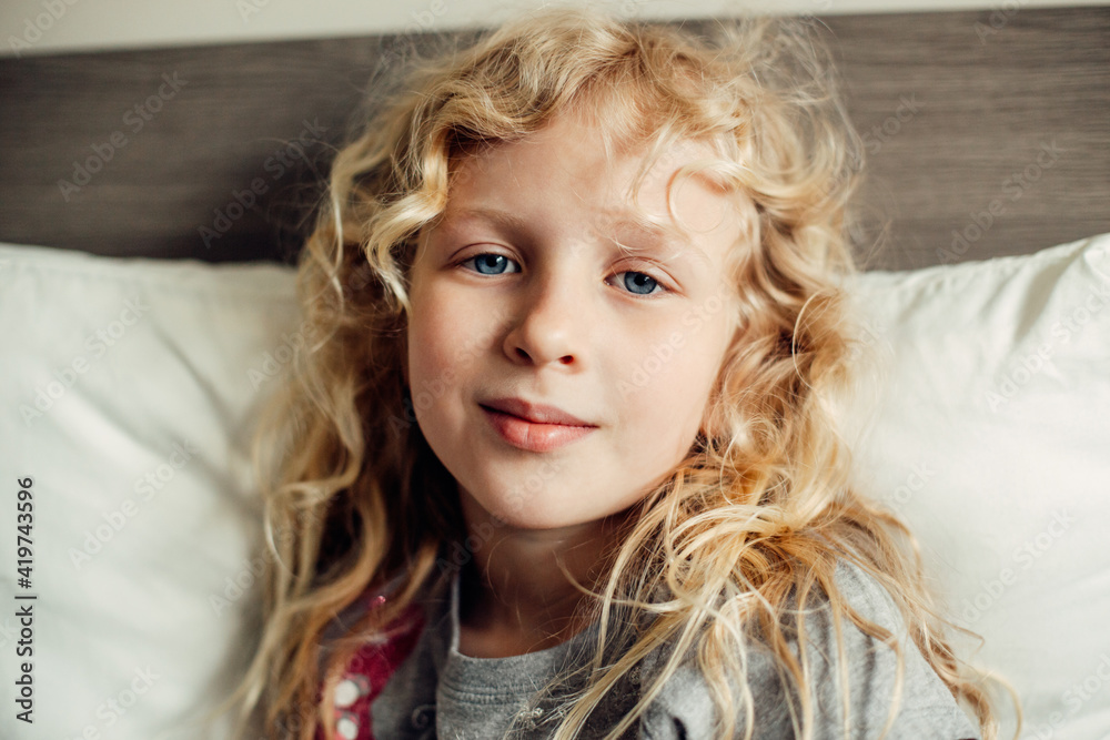 Pretty beautiful Caucasian blonde girl with long messy hair and blue eyes sitting on bed at home looking at camera. Pensive child kid thinking dreaming. Happy authentic childhood lifestyle. Pretty beautiful Caucasian blonde girl with long messy hair and blue eyes sitting on bed at home looking at camera. Pensive child kid thinking dreaming. Happy authentic childhood lifestyle.