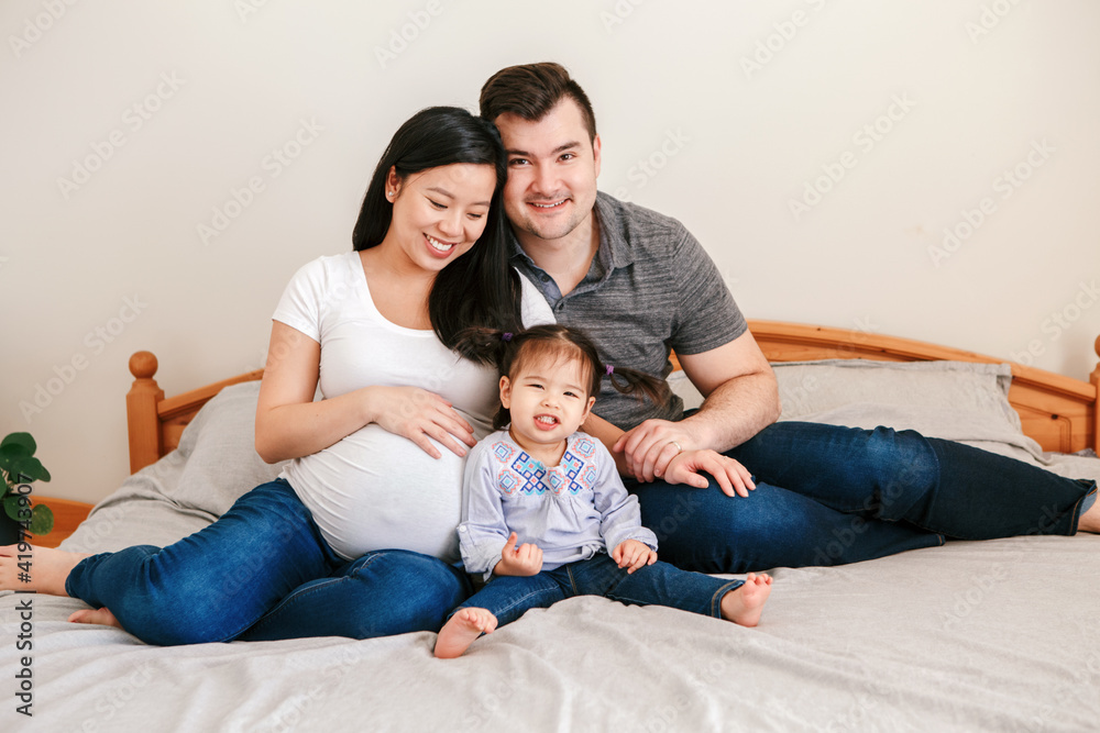 Family Asian Chinese pregnant woman and Caucasian man with mixed race toddler girl sitting on bed at home. Mother, father, baby daughter expecting new family member. Ethnic diversity. Family Asian Chinese pregnant woman and Caucasian man with mixed race toddler girl sitting on bed at home. Mother, father, baby daughter expecting new family member. Ethnic diversity.