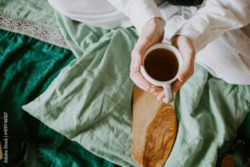 Women's hands hold a hot cup of tea on the bed, linen bedding, country life, relaxation, tranquility, digital detox. High quality photo