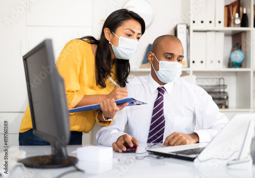 Portrait of businessman with female colleague working in office in medical face masks to prevent spread of viral infection. New life reality