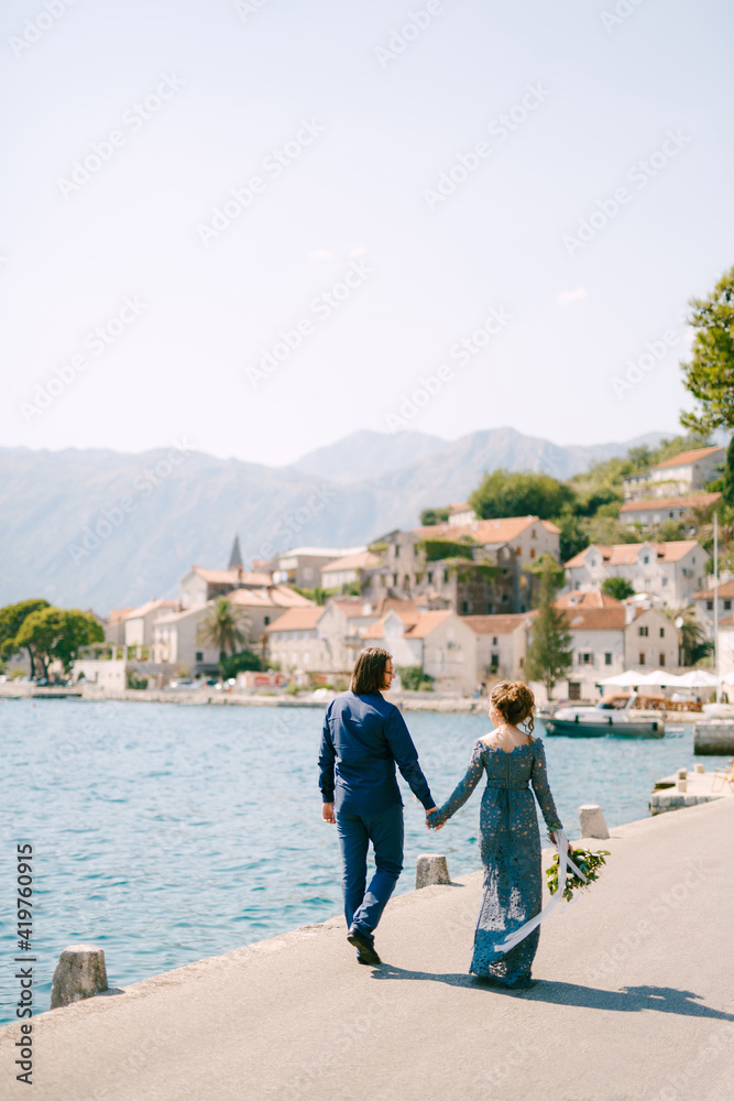 The bride in a stylish blue dress and groom walk along the pier holding hands near the old town of Perast, back view 