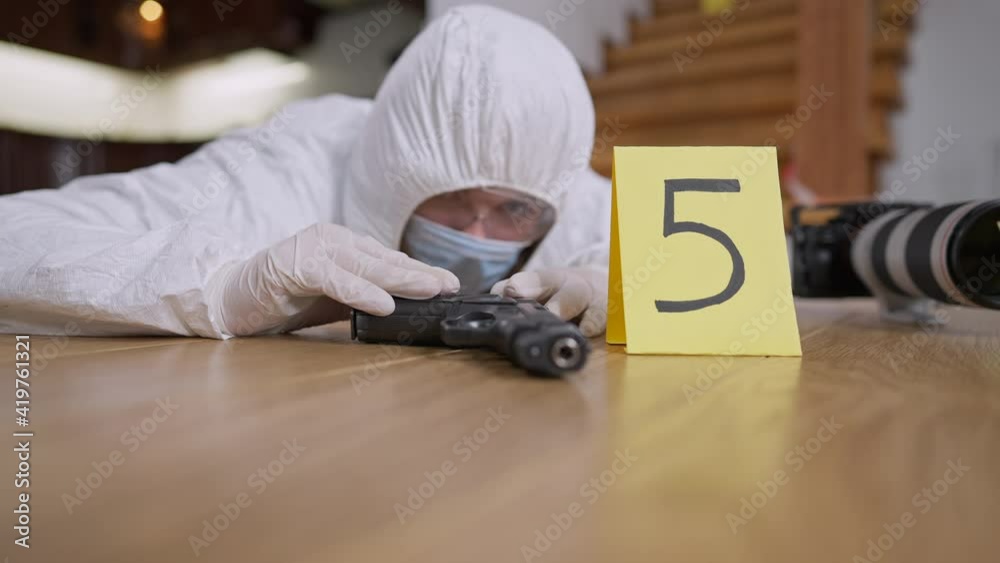 Weapon lying on floor with blurred forensic scientist examining evidence. Focused Caucasian woman working at crime scene indoors. Murder investigation and forensic science concept.