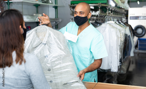 Male employee in protective mask with client in modern laundry, returning clothing after dry cleaning