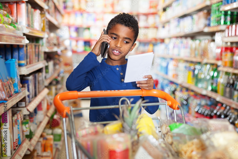 African American preteen boy choosing food goods with shopping list and talking by phone in shop