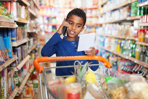 African American preteen boy choosing food goods with shopping list and talking by phone in shop