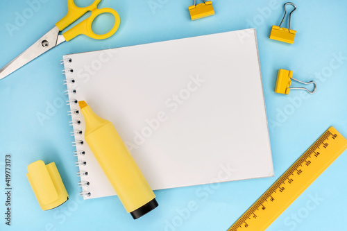 On a blue background among yellow stationery there is a notebook with a blank white sheet. Ruler and scissors with a marker. Template. Education