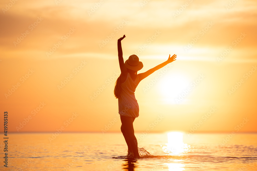 Lady's silhouette stands with raised arms at sunset beach