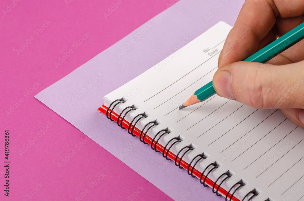 Hand holding a spring-loaded notepad and pencil on a pink and li Hand holding a spring-loaded notepad and pencil on a pink and li