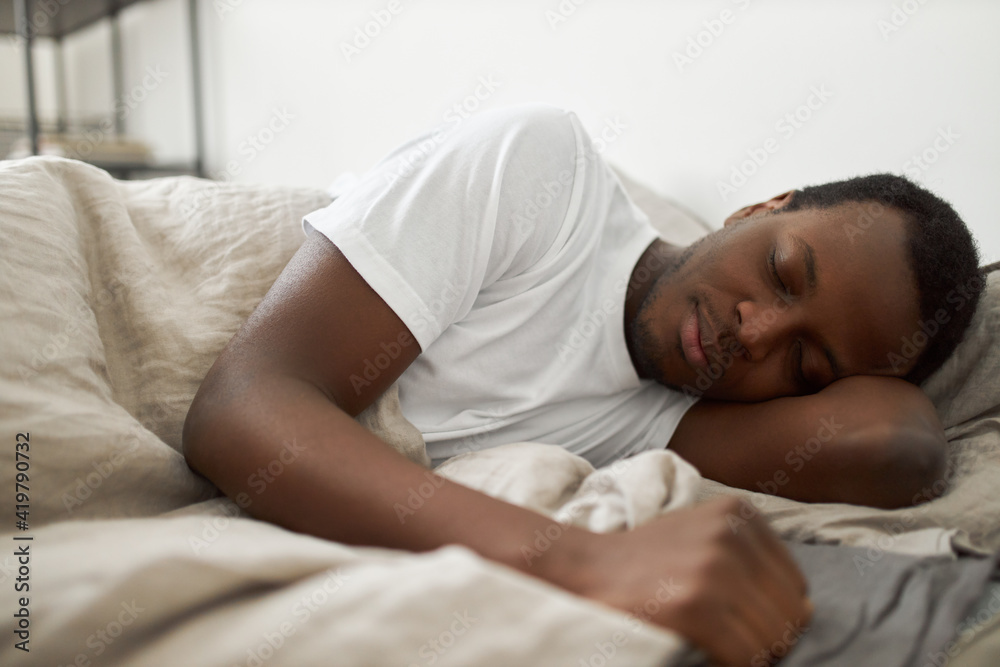 People, bedding, bedtime, rest and relaxation concept. Indoor shot of handsome African American male wearing white t-shirt lying in bed, sleeping peacefully, having pleasant good dream People, bedding, bedtime, rest and relaxation concept. Indoor shot of handsome African American male wearing white t-shirt lying in bed, sleeping peacefully, having pleasant good dream
