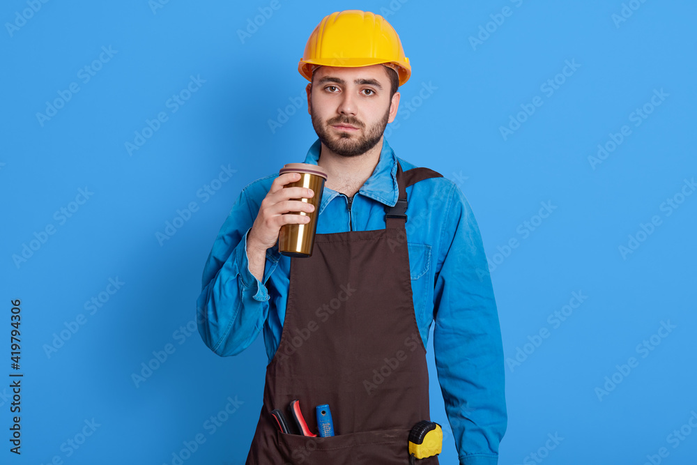 Pleased foreman wears yellow helmet, uniform and brown apron, drinks takeout coffee from thermo mug, looks at camera with tired and exhausted facial expression, posing against blue wall.