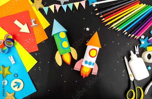 Two space rockets made of colored paper on a black table with colored pencils, colored paper, scissors and glue. The concept of crafts by April 12 is the day of cosmonovics.