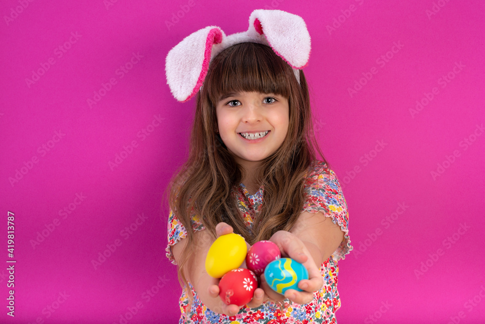 Cute little child girl  smile beaming wearing bunny ears and  standing to holds colored Easter eggs  on pink background