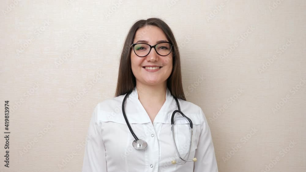 Pretty brunette lady doctor with glasses and stethoscope in white robe smiles to camera standing near beige wall in clinic office closeup Pretty brunette lady doctor with glasses and stethoscope in white robe smiles to camera standing near beige wall in clinic office closeup