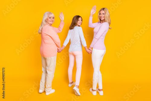 Full size back photo of optimistic three woman grand mom daughter go wave hand wear pastel cloth isolated on yellow background