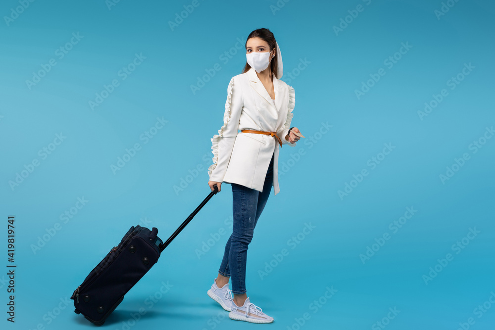 Full-length photo of a woman in a mask with a suitcase ready to fly. Travel, vacation, business trip. Blue background with space. Full-length photo of a woman in a mask with a suitcase ready to fly. Travel, vacation, business trip. Blue background with space.