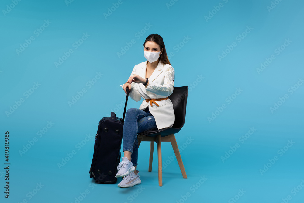 Caucasian girl waiting for departure with a suitcase. Looks at the time on the clock. Business trip. Blue background with simple space.