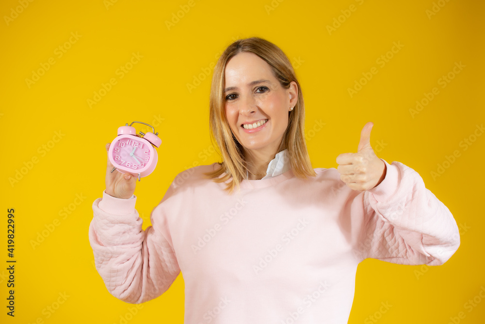Young woman with brown eyes holding alarm clock standing over isolated yellow background very happy with okay sign gesture Young woman with brown eyes holding alarm clock standing over isolated yellow background very happy with okay sign gesture