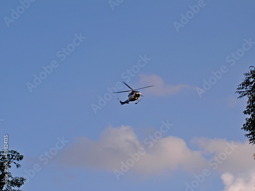 Police helicopter flying in the blue sky during the day, tree branches on the edges, front view